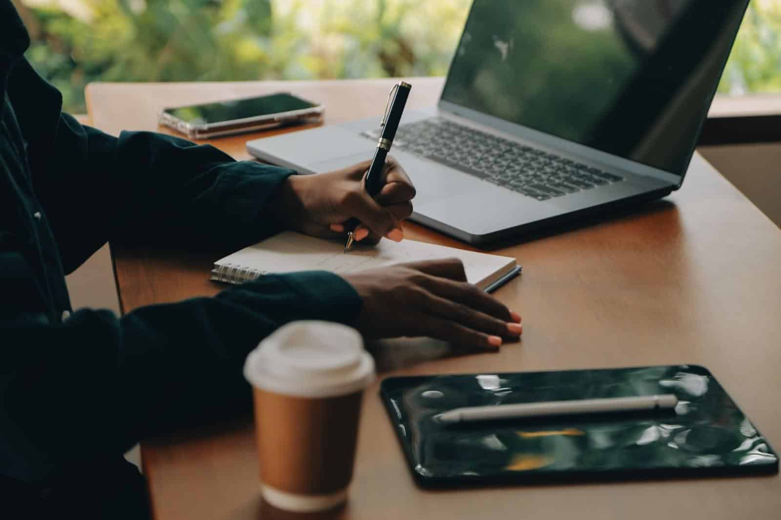 Pessoa escrevendo notas em um caderno ao lado de um laptop e uma xícara de café em uma mesa de madeira.